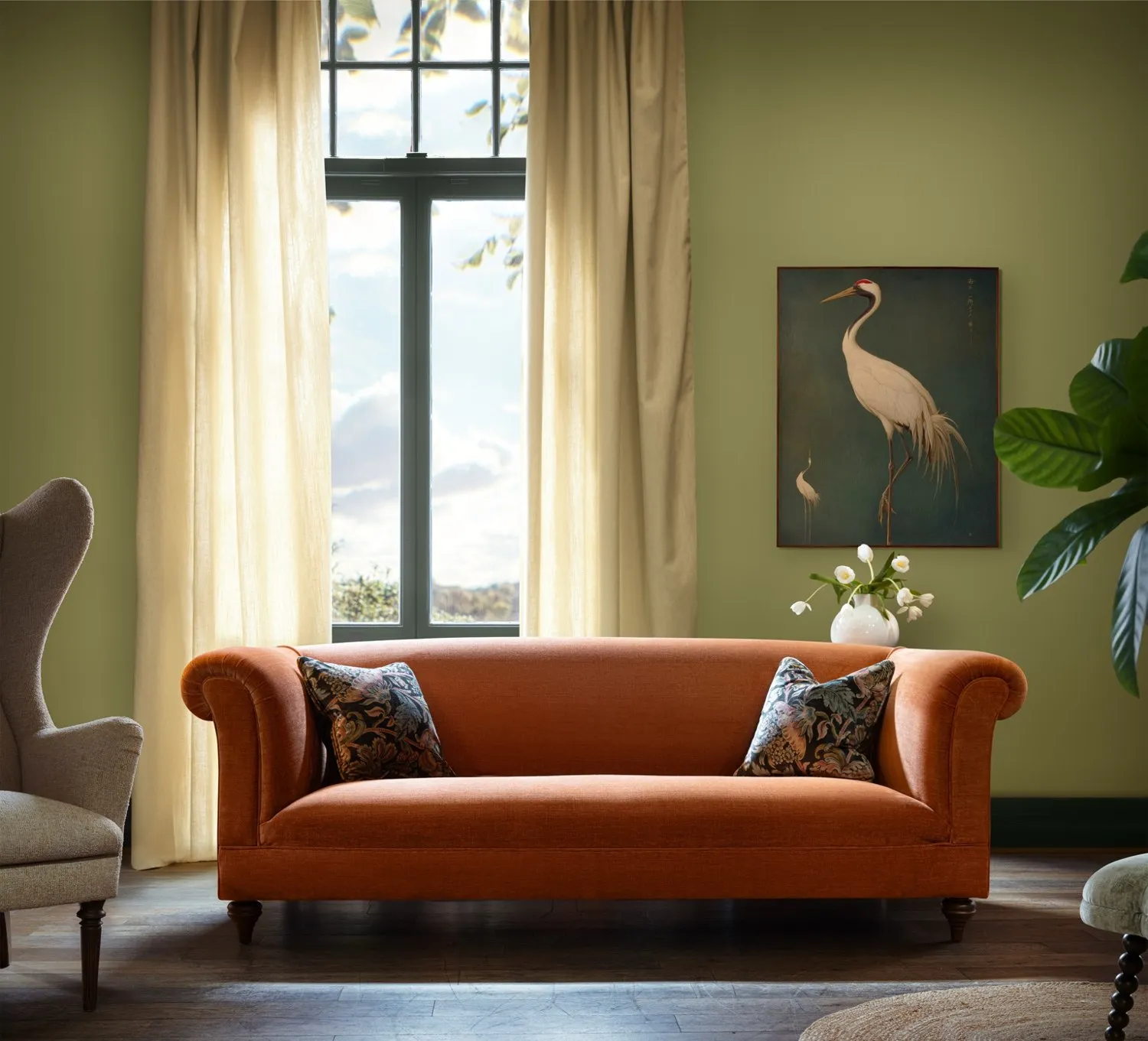 An orange John Sankey sofa in a green room beside a tall window